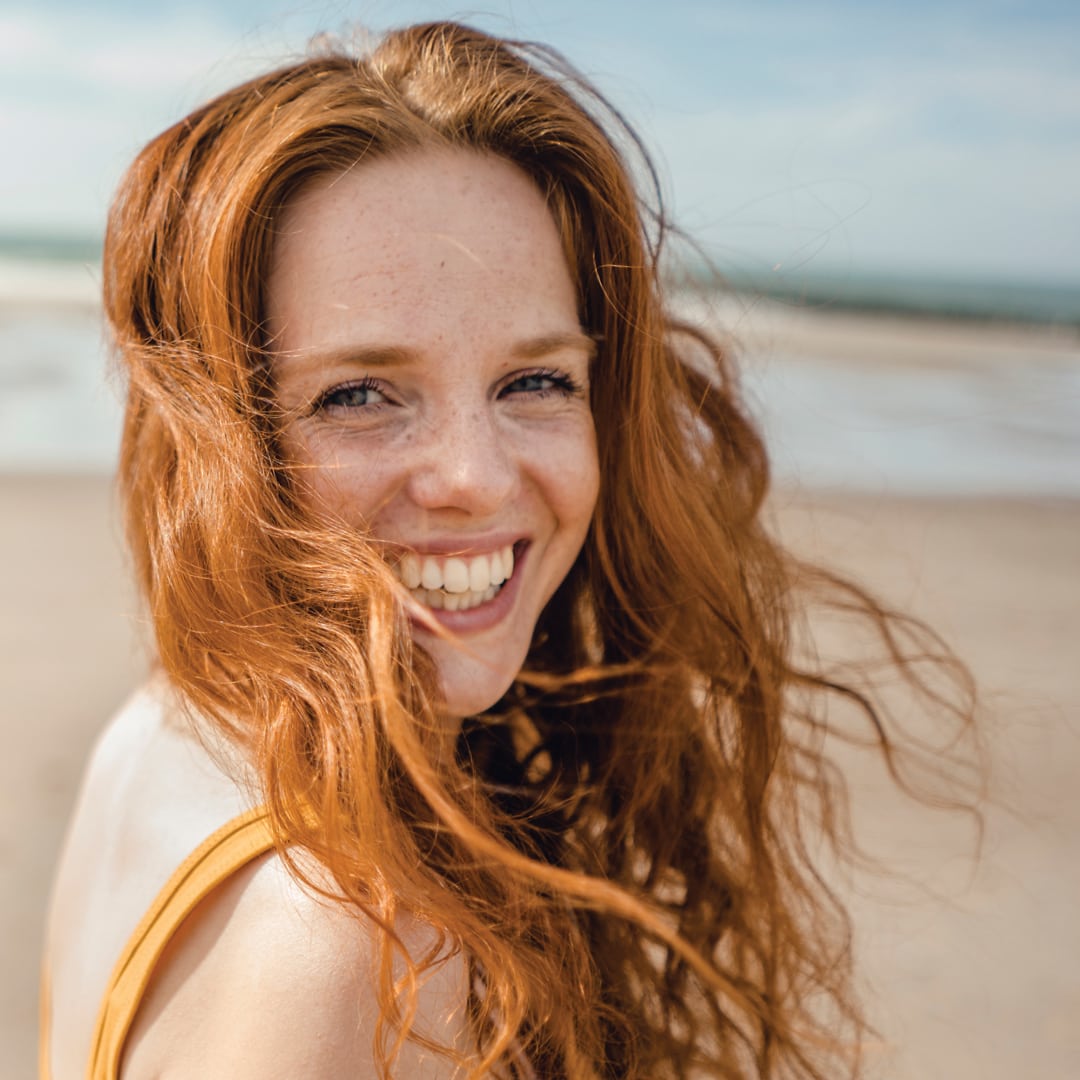 head and shoulders shot of a woman on a beach with rich red hair curly long hair blowing in the wind
