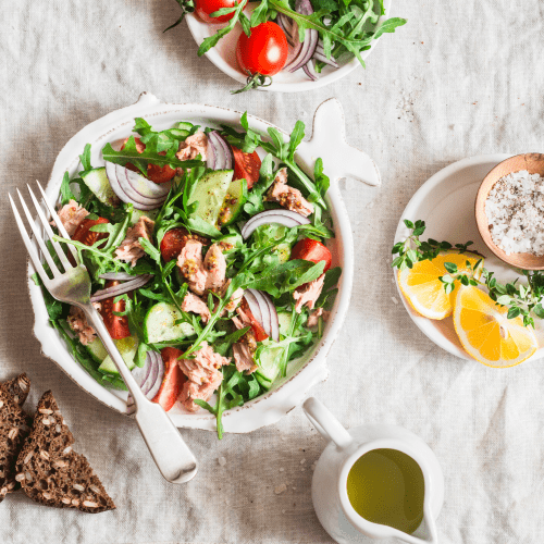 looking down on a big serving of salad. It's very colourful with big pieces of tuna, sliced tomatoes, cucumber and red onion. there is a server of oil dressing beside and a dish of sliced lemons as well