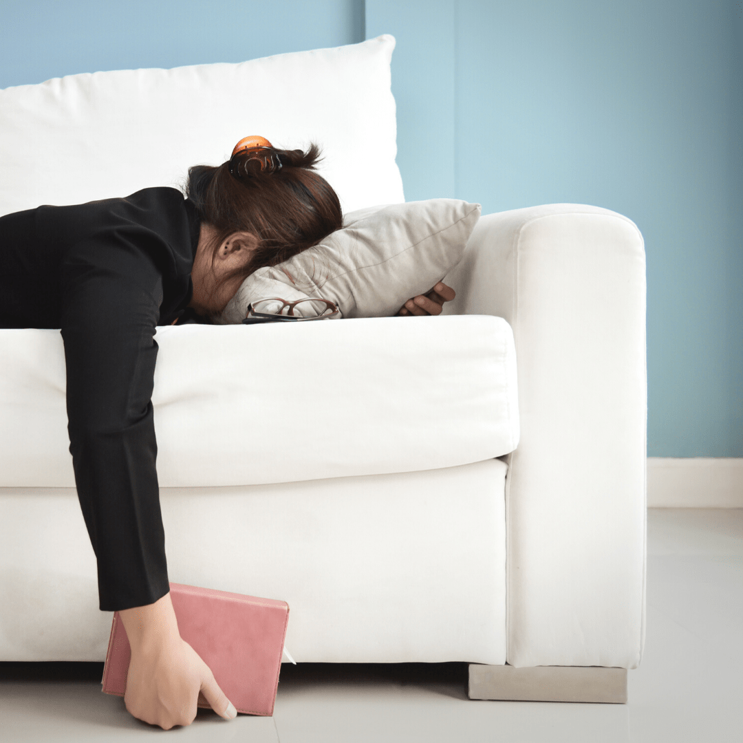 A woman laying face down on a couch. She has a book in her hand but she has fallen asleep.