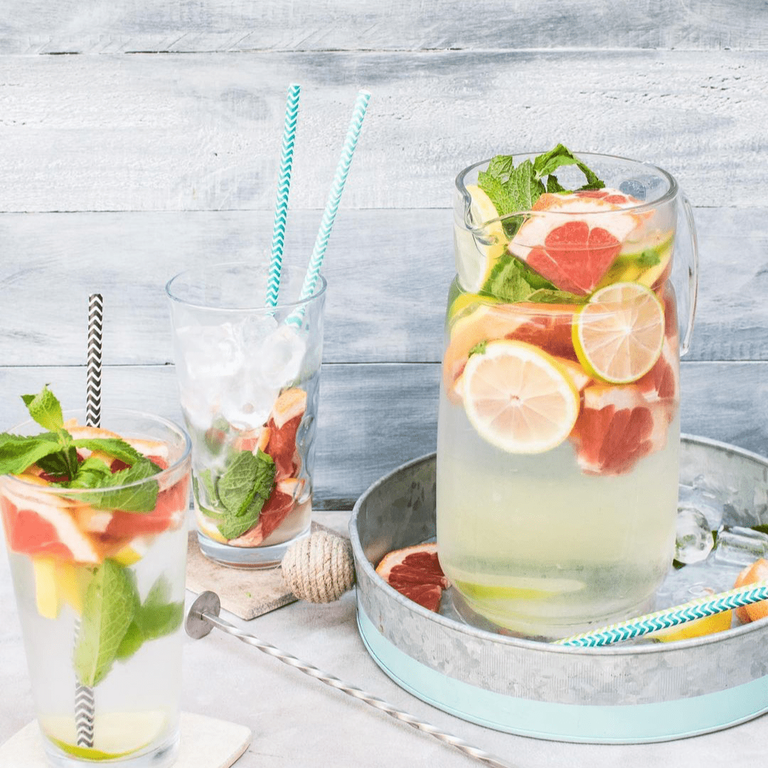 a glass pitcher of water with lots of fruit in it. grapefruits and lemon. There are also mint leaves. Beside it are a couple of glasses with the same mix in them and some ice cubes and stripped straws